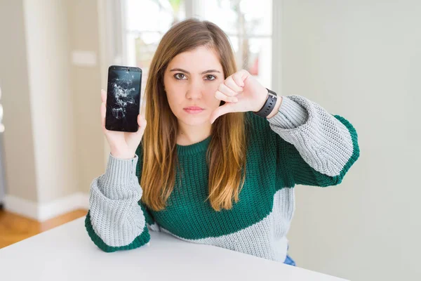 Beautiful young woman holding broken smartphone with angry face, negative sign showing dislike with thumbs down, rejection concept