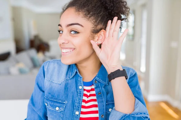 Beautiful young african american woman with afro hair wearing casual denim jacket smiling with hand over ear listening an hearing to rumor or gossip. Deafness concept.