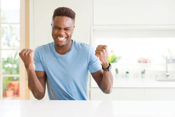 自宅でカジュアルなTシャツを着ているハンサムなアフリカ系アメリカ人男性は 腕を上げ 笑顔を浮かべて 成功のために叫んで勝者のジェスチャーをやって非常に幸せで興奮しています お祝いのコンセプト — ストック写真