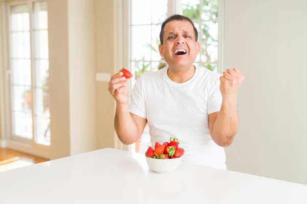 Middle age man eating strawberries at home screaming proud and celebrating victory and success very excited, cheering emotion