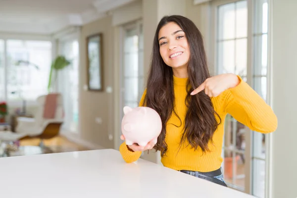 Young woman holding piggy bank with surprise face pointing finger to himself