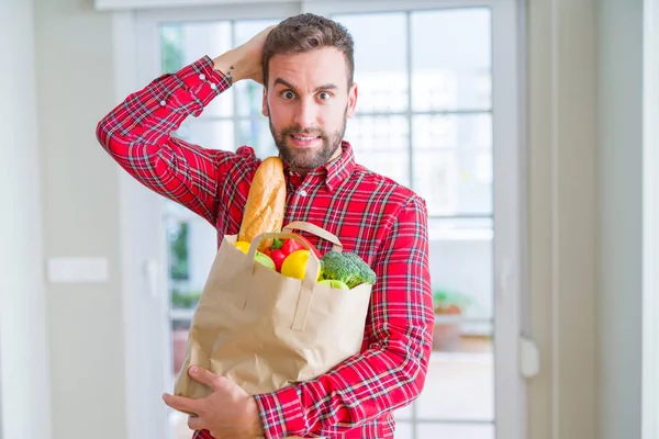 Handsome man holding groceries bag stressed with hand on head, shocked with shame and surprise face, angry and frustrated. Fear and upset for mistake.