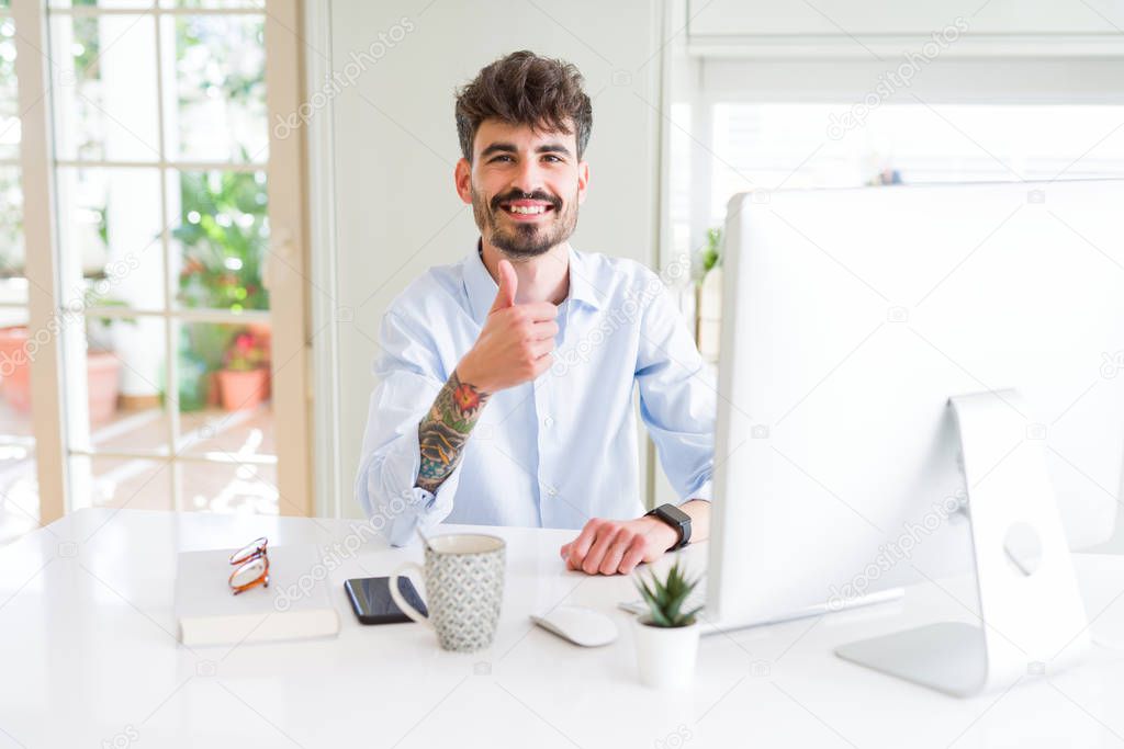 Young business man working using computer doing happy thumbs up gesture with hand. Approving expression looking at the camera with showing success.