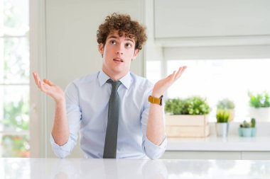 Young business man wearing a tie clueless and confused expression with arms and hands raised. Doubt concept. clipart