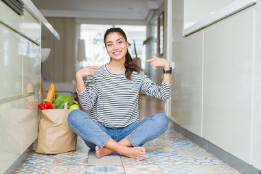 Mutfak zemininde oturan genç bir kadın, elinde bir torba dolusu taze erzakla kendine güvenen, yüzünde bir gülümsemeyle, gururlu ve mutlu bir şekilde kendini gösteren bir kadın..