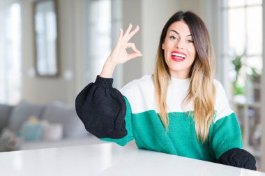 Genç güzel kadın giyiyor kış kazak evde el ve parmaklar ok işaretiyle yapıyor olumlu gülümseyerek. Başarılı ifade.