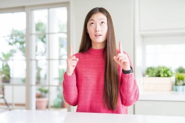 Beyaz masada pembe kazak giyen güzel Asyalı kadın şaşırttı ve yukarı bakarak ve parmakları ile işaret ve kolları kaldırdı şaşırttı.