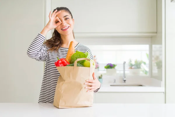 Young woman holding paper bag full of groceries with happy face smiling doing ok sign with hand on eye looking through fingers