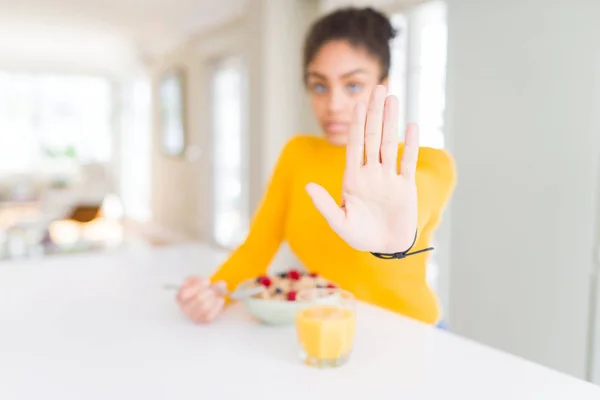 Young african american girl eating healthy cereals for breakfast with open hand doing stop sign with serious and confident expression, defense gesture