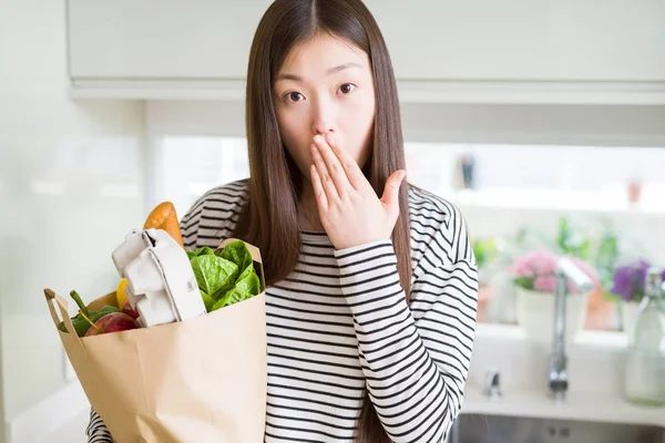 Beautiful Asian woman holding paper bag of fresh groceries cover mouth with hand shocked with shame for mistake, expression of fear, scared in silence, secret concept