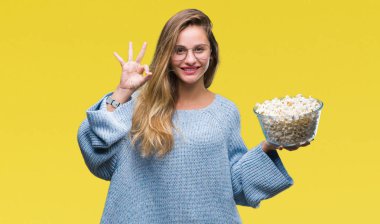 Genç güzel sarışın kadın parmakları, mükemmel sembolü olan ok işareti yapıyor izole arka plan üzerinde patlamış mısır yemek