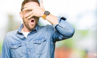 Şok amorti eden gözlük takan yakışıklı adam, yüzünü ve gözlerini eliyle örterek, utanmış ifadeyle parmaklarından bakıyor.