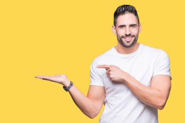 Hayret ve el ile sunulması ve parmak ile işaret ederken kameraya gülümseyen izole arka plan üzerinde rahat beyaz t-shirt giyen genç adam.