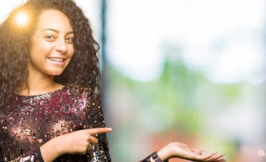 El ile sunulması ve parmak ile işaret ederken gece parti giyen kıvırcık saçlı genç güzel kız elbise hayret ve kameraya gülümseyen.