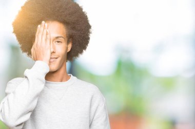 El yüz ve sürpriz duygu kendinden emin gülümseme ile tek gözüyle kapsayan sportif sweatshirt giyen afro saçlı genç Afro-Amerikan adam.