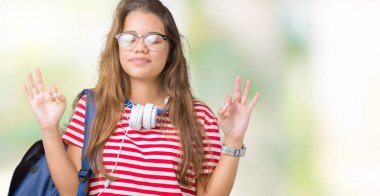 Kulaklık ve sırt çantası izole arka plan üzerinde giyen genç güzel esmer öğrenci kadın dinlenmek ve gülümseyen gözlerle kapalı parmak jestle meditasyon yapıyor. Yoga kavramı.