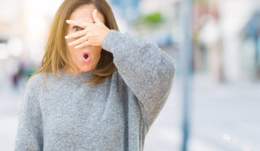 Izole arka plan üzerinde kış kazak giyen güzel orta yaş kadın şok yüz ve gözleri el ile kaplayan gözetleme, utanmış ifade ile parmaklarından bakarak.