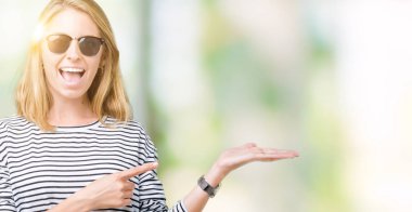 Güzel genç kadın hayrete ve el ile sunulması ve parmak ile işaret ederken kameraya gülümseyen izole arka plan üzerinde güneş gözlüğü takıyor.