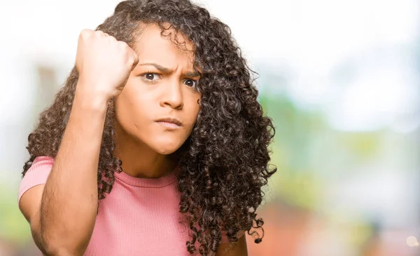 Young beautiful woman with curly hair wearing pink t-shirt angry and mad raising fist frustrated and furious while shouting with anger. Rage and aggressive concept.