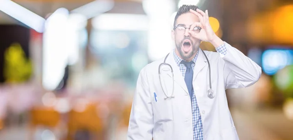 Handsome young doctor man over isolated background doing ok gesture shocked with surprised face, eye looking through fingers. Unbelieving expression.