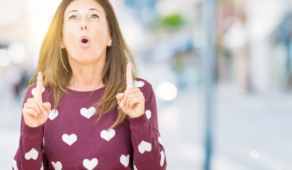 Beautiful middle age woman wearing hearts sweater over isolated background amazed and surprised looking up and pointing with fingers and raised arms.