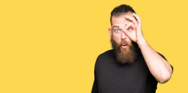 Sürpriz yüz, göz parmaklarının arasından seyir ile ok jest yapıyor rahat t-shirt giyen genç sarışın adam şok. İmansız ifade.