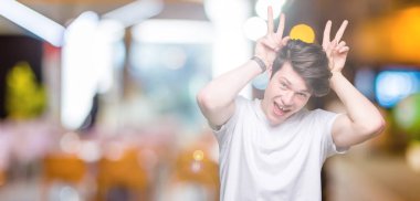 Neşeli gülümseyen izole arka plan tavşan kulakları, komik ve çılgın baş parmaklar ile poz üzerinde rahat beyaz t-shirt giyen genç yakışıklı adam