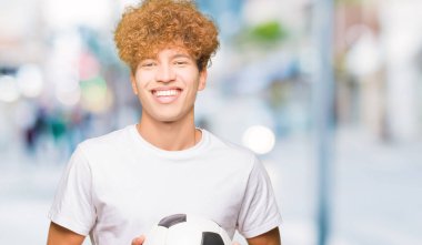 Genç yakışıklı adam ayakta mutlu bir yüz ile futbol topu tutan ve dişleri gösteren kendinden emin bir gülümseme ile gülümseyerek