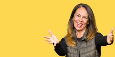 Sarılmak için kollarını açarak gülümseyerek kameraya bakarak kış yelek giyen güzel orta yaş kadın. Neşeli ifade kucaklayan mutluluk.