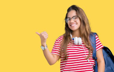 Kulaklıklar ve sırt çantası arıyorum ve başparmak yanında işaret mutlu yüz ile gülümseyen izole arka plan üzerinde giyen genç güzel esmer öğrenci kadın.