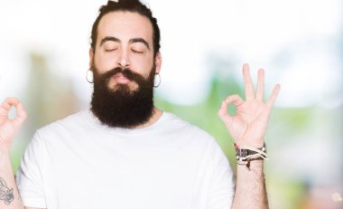 Uzun saç ve sakal casual beyaz t-shirt giyen genç hipster adamla rahatla ve gülümseyen gözlerle kapalı parmak jestle meditasyon yapıyor. Yoga kavramı.