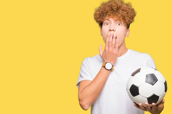 Young handsome man holding soccer football ball cover mouth with hand shocked with shame for mistake, expression of fear, scared in silence, secret concept