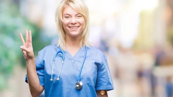 年轻美丽的金发医生妇女穿医疗制服在孤立的背景显示和指向与手指数字三同时微笑自信和快乐 — 图库照片