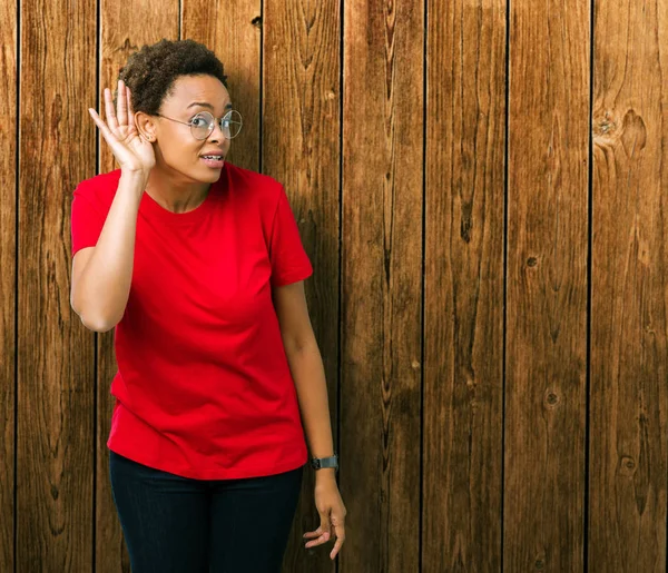 Beautiful young african american woman wearing glasses over isolated background smiling with hand over ear listening an hearing to rumor or gossip. Deafness concept.