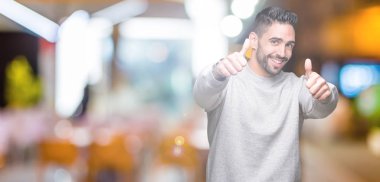 El ile pozitif jest yapıyor onaylama izole arka plan üzerinde sweatshirt giyen genç yakışıklı adam başparmak gülümseyen ve başarı için mutlu. Kamera, kazanan jest arıyorsunuz.