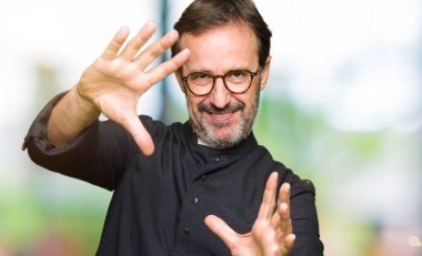 Orta yaş rahip adam katolik cüppe giyen Gülen elleri avuç içi ve parmaklar kullanarak çerçeve yapıyor, kamera perspektifi