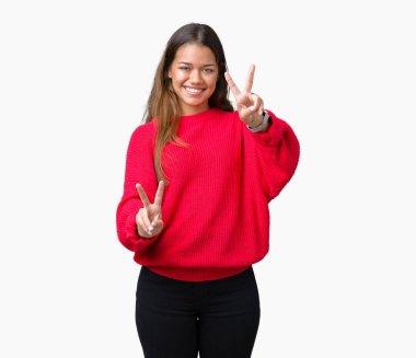 Zafer işareti yapıyor parmak gösterilen kamera seyir gülümseyen izole arka plan üzerinde kırmızı kış kazak giyen genç güzel esmer kadın. İki numaralı.