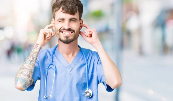 Young handsome nurse man wearing surgeon uniform over isolated background covering ears with fingers with annoyed expression for the noise of loud music. Deaf concept.