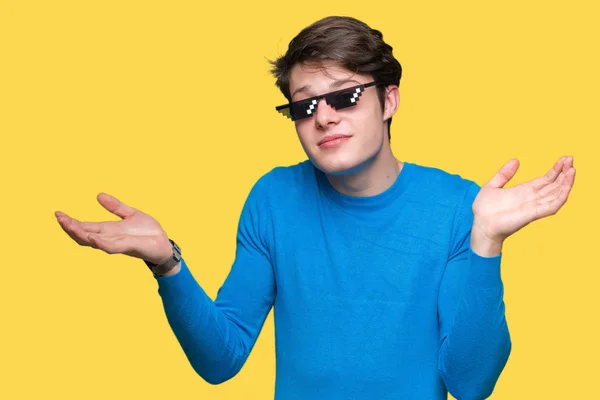 Young man wearing funny thug life glasses over isolated background clueless and confused expression with arms and hands raised. Doubt concept.
