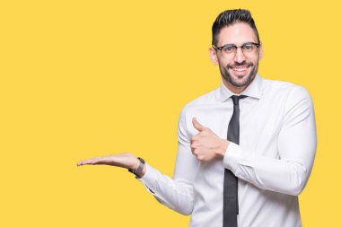 Üzerinde gözlük takan genç yakışıklı iş adamı arka plan gösteren palm el ve iş tamam jestle başparmak yukarıya, mutlu ve neşeli gülümseyen izole