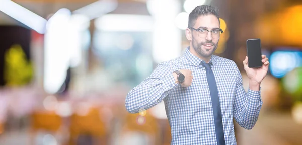 Young business man showing smartphone screen over isolated background with surprise face pointing finger to himself