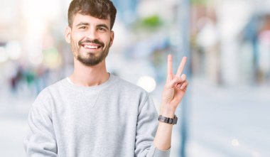 Mutlu yüz zafer işareti yaparken kameraya göz kırpıyor ile gülümseyen izole arka plan üzerinde sweatshirt giyen genç yakışıklı adam. İki numaralı.