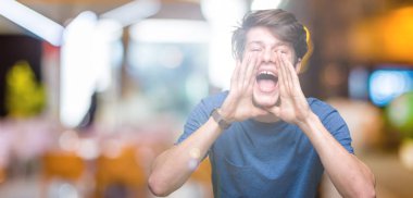 Kızgın ile yüksek sesle bağırarak izole arka plan üzerinde mavi t-shirt giyen genç yakışıklı adam ağzına eller