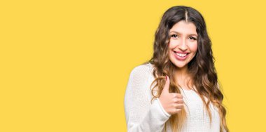 Beyaz kazak giyen genç güzel kadın eliyle jest kadar mutlu başparmak yapıyor. Başarı gösteren kameraya bakarak ifadeyi onaylama.