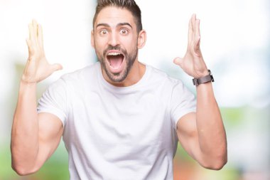 Üzerinde rahat beyaz t-shirt giyen genç adam deli kutluyor arka plan izole ve başarı için kaldırdı kollarını açarak hayrete ve heyecanlı çığlık gözleri açık. Kazanan kavramı