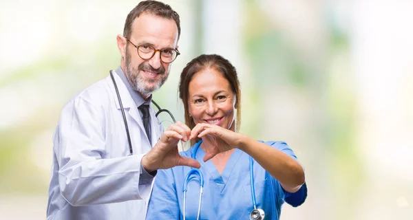 Middle age hispanic doctors partners couple wearing medical uniform over isolated background smiling in love showing heart symbol and shape with hands. Romantic concept.