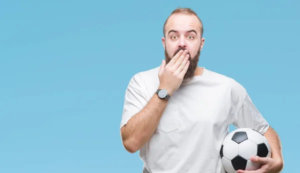 Young caucasian hipster man holding soccer football ball over isolated background cover mouth with hand shocked with shame for mistake, expression of fear, scared in silence, secret concept