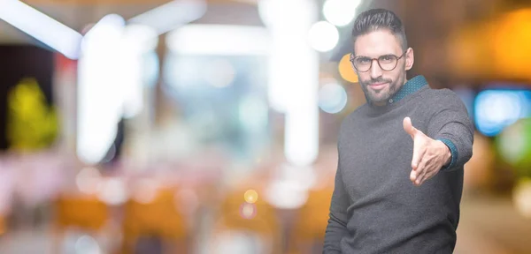 Young handsome man wearing glasses over isolated background smiling friendly offering handshake as greeting and welcoming. Successful business.