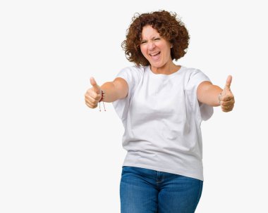 El ile pozitif jest yapıyor onaylama izole arka plan üzerinde beyaz t-shirt giyen kadın güzel orta ager kıdemli başparmak gülümseyen ve başarı için mutlu. Kamera, kazanan jest arıyorsunuz.