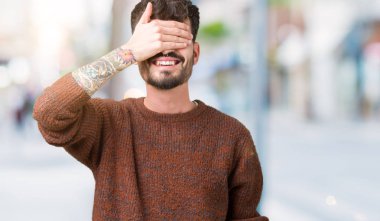 Genç yakışıklı adam gülümsüyor ve gözleri için sürpriz kapsayan yüz el ile gülüyor izole arka plan üzerinde kış kazak giyiyor. Kör kavramı.